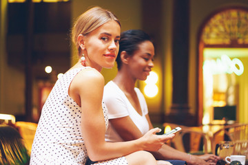 Two female friends are discussing fashion news while drinking coffee in a modern city restaurant. A blonde model look girl with a smartphone is looking at camera while sitting with a friend in a cafe.