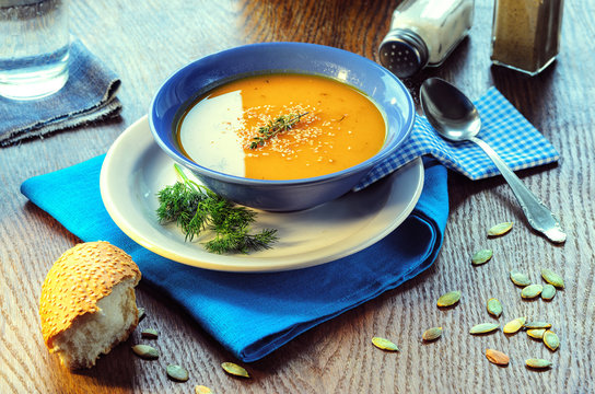 Pumpkin cream-soup. Hot pumpkin soup with toasted sesame seeds in a blue bowl on a wooden table. Still life with a bowl of soup, spoon, pumpkin seeds and slice of baguette