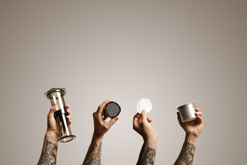 Empty clear aeropress, filter cap, two paper filters and steel travel cup held up in the air by four hands against white background Alternative coffee brewing commercial