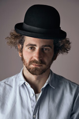 Fototapeta na wymiar Portrait of a sad looking young bearded jewish man with curly hair wearing a funny black bowler hat and light denim button up t-shirt isolated on light gray.