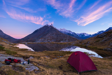 Camping with tent near high altitude lake on the Alps. Reflection of snowcapped mountain range and scenic colorful sky at sunset. Adventure and exploration.