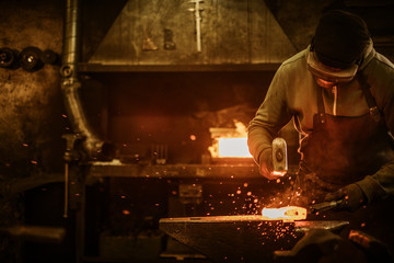 The blacksmith forging the molten metal on the anvil in smithy - obrazy, fototapety, plakaty