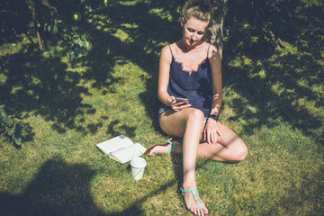 Summer sunny day,young woman with blond hair,wearing blue top and denim shorts,sitting on the lawn and surf the Internet on digital gadget.On hand smartwatch.Nearby are notebook,pen and cup of coffee.