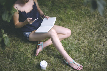 Summer day,girl in blue top and short shorts sitting in garden under tree on green grass and using gadget,on knees is notebook,next on lawn is cup of coffee.Woman surfing Internet on smartphone.