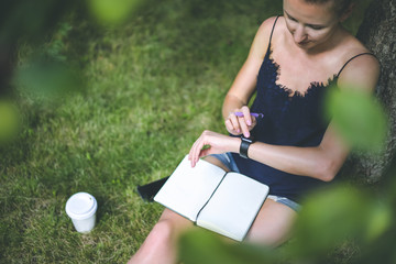 Girl sitting in garden on lawn under tree and uses smartwatch.Woman wearing blue top and short denim shorts,pen in hand,is on her lap lying notebook,near smartphone and cup of coffee.Girl using gadget