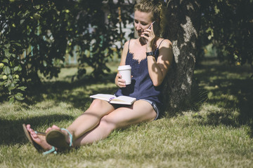 Summer day,girl with blond hair talking on cell phone while sitting on grass under the apple tree. In her hand she holds a cup of coffee on her lap lying notebook.Young woman wearing a top and shorts.