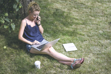 Summer day,woman with blond hair,wearing blue top and denim shorts sitting in garden under tree and talking on phone while using laptop.Nearby on grass notebook and cup of coffee.Girl using gadget.