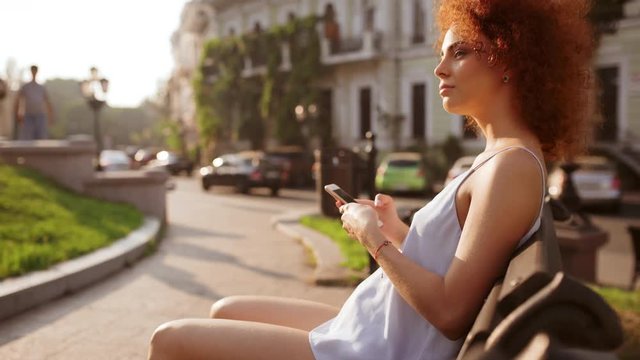 Attractive readhead girl resting looking at phone sitting on bench outside