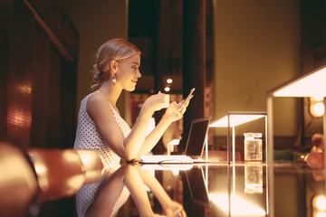 Young model look blonde woman is reading messages on her mobile phone during coffee break in a coffee shop. An entrepreneur female is preparing to the meeting while sitting at the table with laptop.