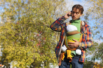 father with baby in sling talking on mobile phone and looking at