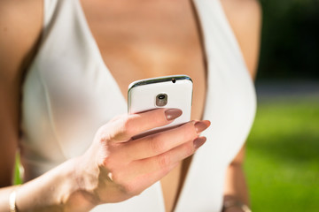 close up photo of young woman with open decolletage reads message to mobile phone in green summer park