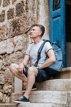 Stylish tourist. Man dressed in a white shirt and blue shorts with a blue backpack over his shoulder. Sitting on the steps of an old European city
