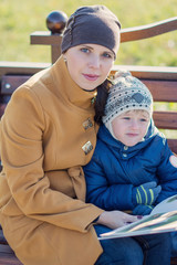 Young woman and little boy sitting on the bench in autumn Park and reading book. Family enjoying time together. Mother teaches son