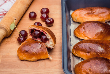 Oven fresh home made baked patties stuffed with cherry. Delicious pastries on a baking sheet, surrounded by cherry, napkin and rolling pin.