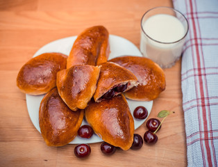 Oven fresh home made baked patties stuffed with cherry. Glass of milk. Delicious pastries on a baking sheet, surrounded by cherry, napkin and rolling pin.