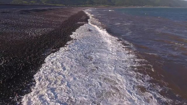 Low aerial view flying along waves washing up onto a pebbled beach.