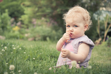 Cute baby girl sitting in grass among blossoming flowers in summer park with copy space
