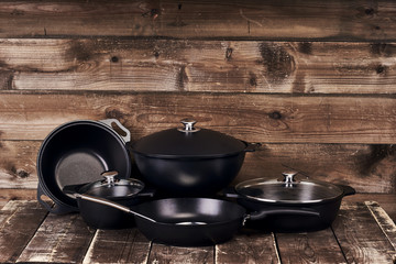 Different black pots, pans and stainless steel laddles on old grunge wooden table against wood wall...
