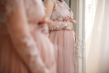 Pregnant woman in a pink dress holding hands on her belly and looks at himself in the mirror. 