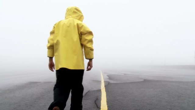 Model released man wearing yellow rain jacket walks away on road during stormy day.