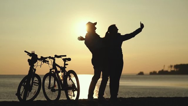 Silhouettes of two cyclists at sunset. The pair finished the bike ride on the river and do selfi on a beautiful background. Couple in love. Unusual date. hotograph on a mobile phone