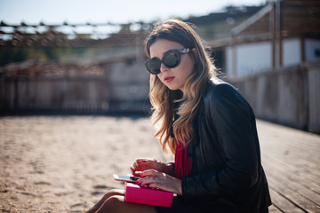 Young beautiful girl in dark clothes and sunglasses sitting on an empty beach and holding a pink box