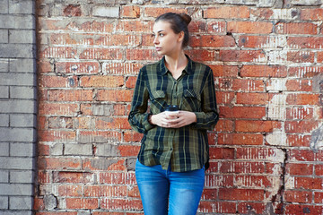 Fototapeta na wymiar A brunette woman in standing disposable coffee cup on the background the red brick wall