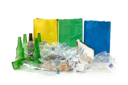 Pile of paper waste, used plastic and glass bottles are on the white background. Empty yellow, green and blue bags are standing behind them.
