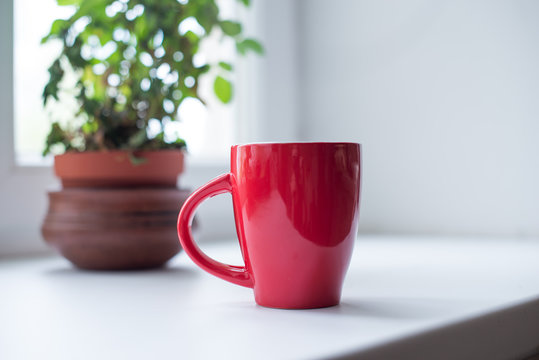 Red mug with tea or coffee on white table by the window