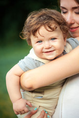 Young beautiful mother hugging her little toddler son against green grass. Happy woman with her baby boy on a summer sunny day. Family walking on the meadow.