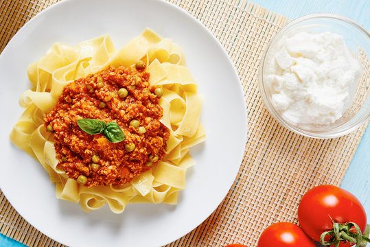 Tagliatelle with Bolognese sauce in white dish surrounded by fresh tomatoes and ricotta cheese on blue table