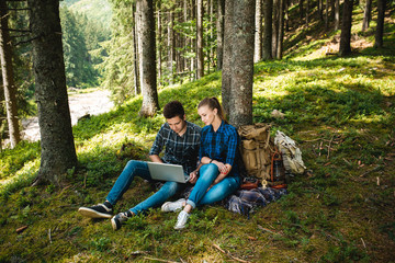 Fototapeta premium A guy and a girl tourists relax in the mountain forest, sitting and looking at laptop