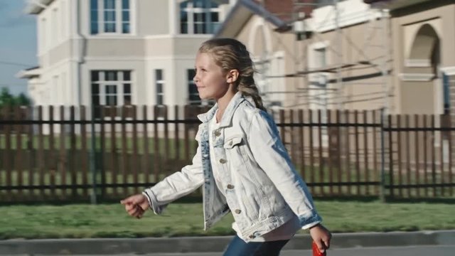 Slow motion tracking of little girl in jeans jacket running through street in countryside on fine summer day