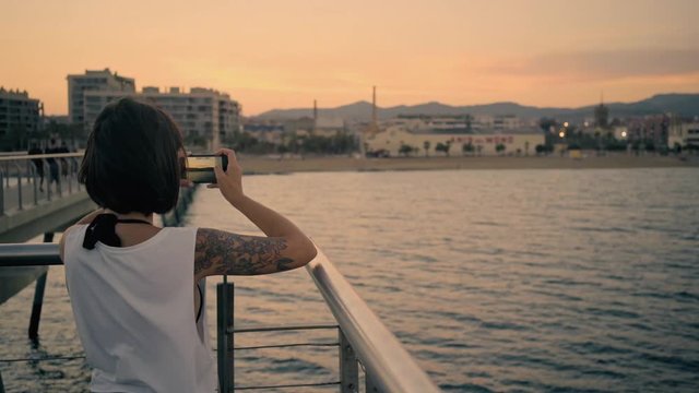 Attractive brunette girl with tattoo takes photo or video of sunset on her smartphone while standing on sea pier in front of city beach Wind sways her hair repeatedly Looped cinemagraph.