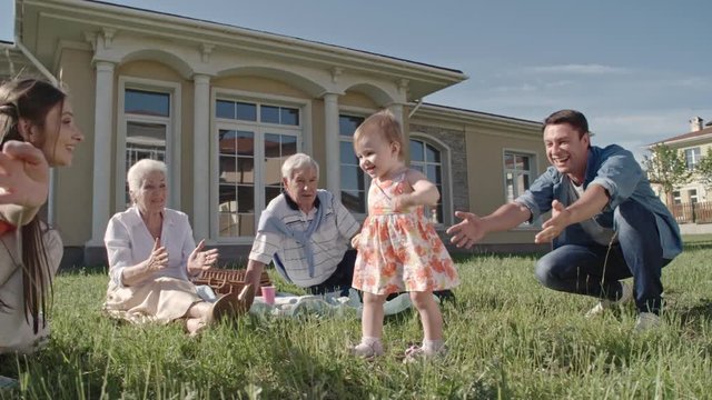 Slow motion of blond toddler girl in dress walking from father to mother, then hugging her and laughing 