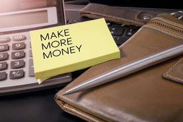 Text Make More Money on yellow note paper calculator pen and leather wallet on laptop.