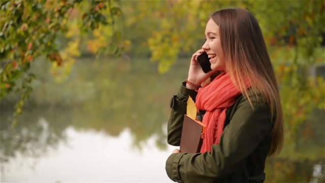 Young female talking on cellphone in the autumn garden near lake, beautiful girl laughing, public park background