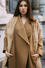 Beautiful girl with hair and make-up, walking around the city / standing near a big blue door next to the columns. Dressed in a long warm beige coat and a fur scarf. Street style.