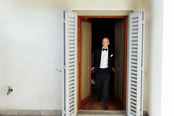 Bald-headed man in black tuxedo walks from the glass doors