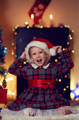 Portrait of happy smiling girl in Santa hat and festive dress sitting on the floor in decorated room over garland lights background. Christmas wish and miracle concept.  