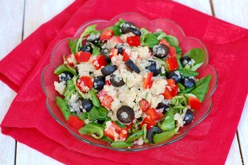 Fresh healthy vegan salad with quinoa, corn salad, black olives, red pepper and olive oil in glass bowl on red cloth on white wooden background