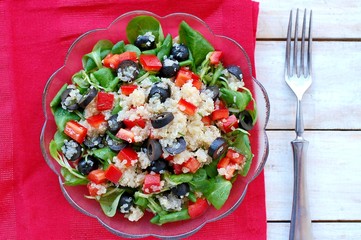 Fresh healthy vegan salad with quinoa, corn salad, black olives, red pepper and olive oil in glass bowl on red cloth on white wooden background