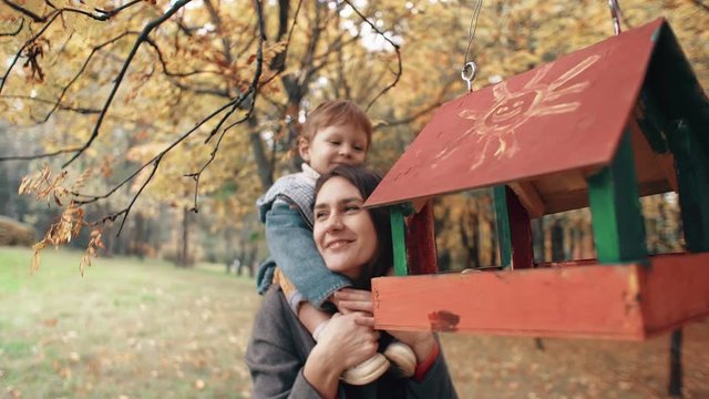 young mother holds on shoulders cute little boy, little boy hugs kisses her near bird feeders in amazing autumn park 4k