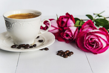 Beautiful white cup of coffee, coffee beans and purple roses on white wooden background, upper view close-up