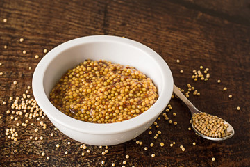 Mustard.  Cooked mustard in a white cup and mustard seeds in a spoon on a brown wooden table.