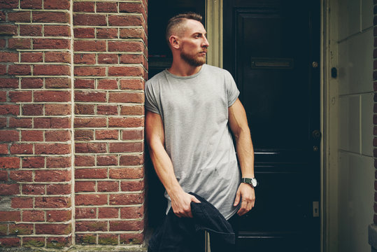 Young bearded hipster wearing a blank gray t-shirt is looking aside while leaning on a brick wall on the street. A photo of a male model in a blank gray t-shirt with empty place for the logo or design