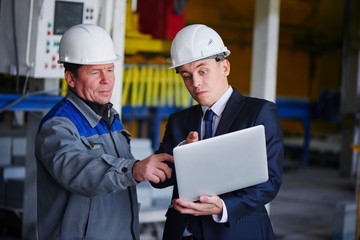 The man in the suit and the helmet holds the portable computer and shows up on the screen to the worker in overalls in an industrial building