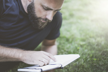 Summer day. Front view of a young bearded man, wearing a black T-shirt, lying on the lawn and reading a book.Selective focus, film effect, blurred background.