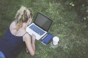 Summer,rear view and top view,girl with blond curly hair lying on grass and using laptop with blank screen.Next to computer is black notebook,pen,smartphone,wired headphones and a white coffee cup.
