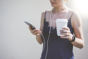 Summer sunny day,front view,girl listening to music with headphones and using smartphone,in hand holding white cup of coffee.On women's hand of smartwatch.In background a white wall.Girl using gadget.